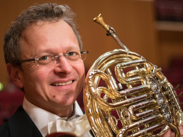 Ein Hornist mit grauen Locken und Brille, im Konzertanzug, hält sein Instrument in die Kamera.
