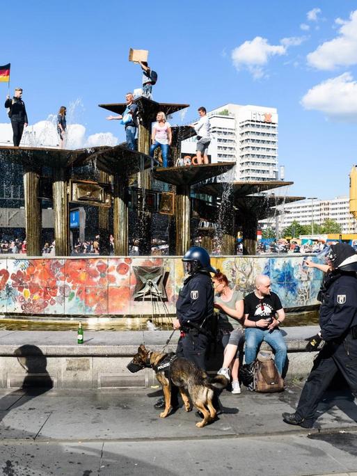Vor dem Brunnen auf dem Alexanderplatz in Berlin gehen Polizisten. Dahinter haben Demonstranten den Brunnen bestiegen.