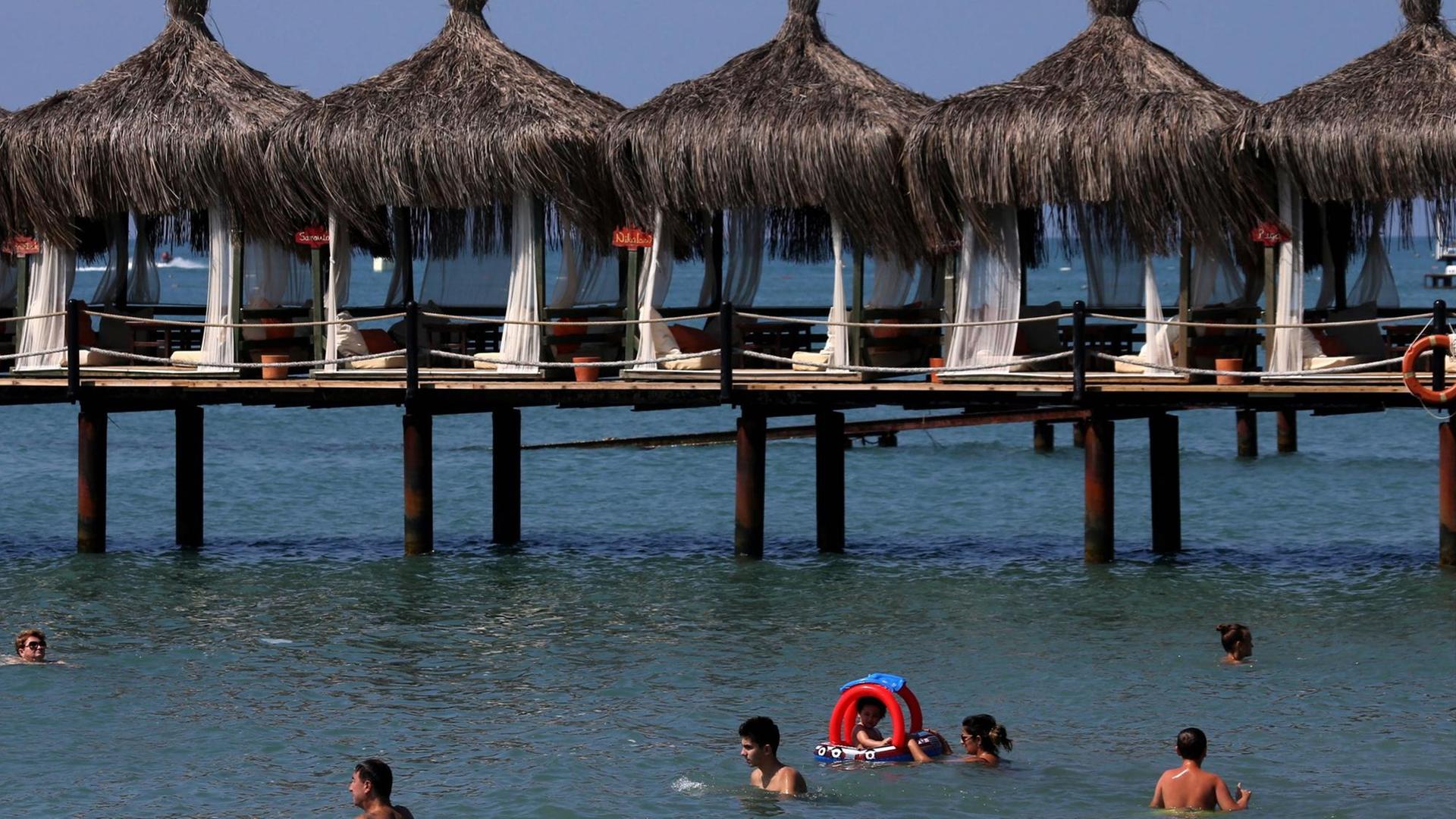 Urlaubsgäste schwimmen im Meer am Strand von Antalya. Dahinter einer Anlage mit Sonnenschirmen und Liegen, die auf Holzpfälen über dem Wasser aufgestellt ist.