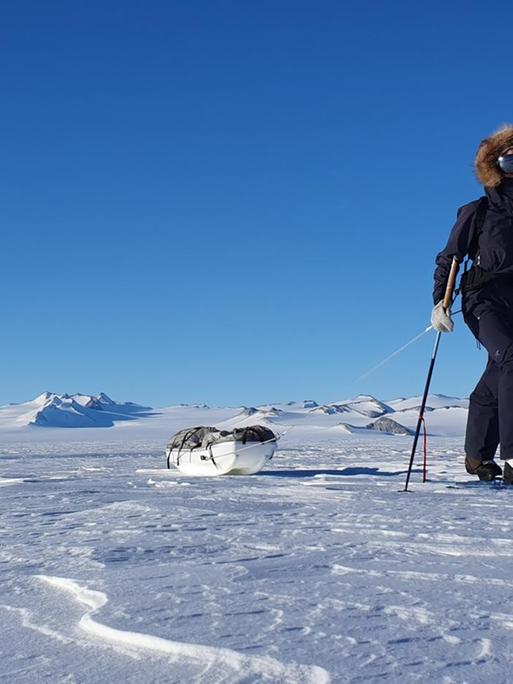 Eine Frau ist alleine auf Langlauf-Skiern mit umgeschnalltem Schlitten, auf dem ein Ruckack liegt, zu sehen.