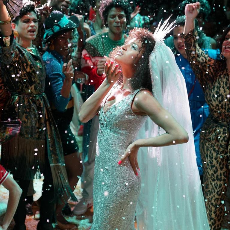 US-Schauspielerin Indya Moore steht in der Rolle der Transfrau Angel in einem weißen Glitzerkleid mit Haube umringt von anderen Tänzern auf dem Parkett, dabei regnet es Glitter