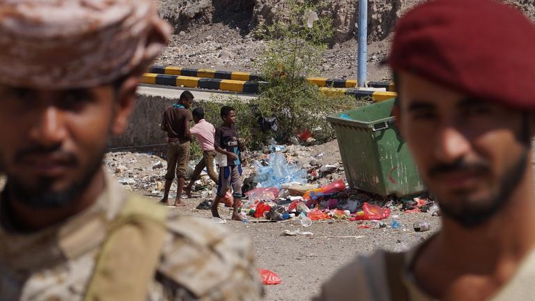 Zwei Männer stehen im Vordergrund. Im Hintergrund spielen Kinder vor einem Müllberg.
