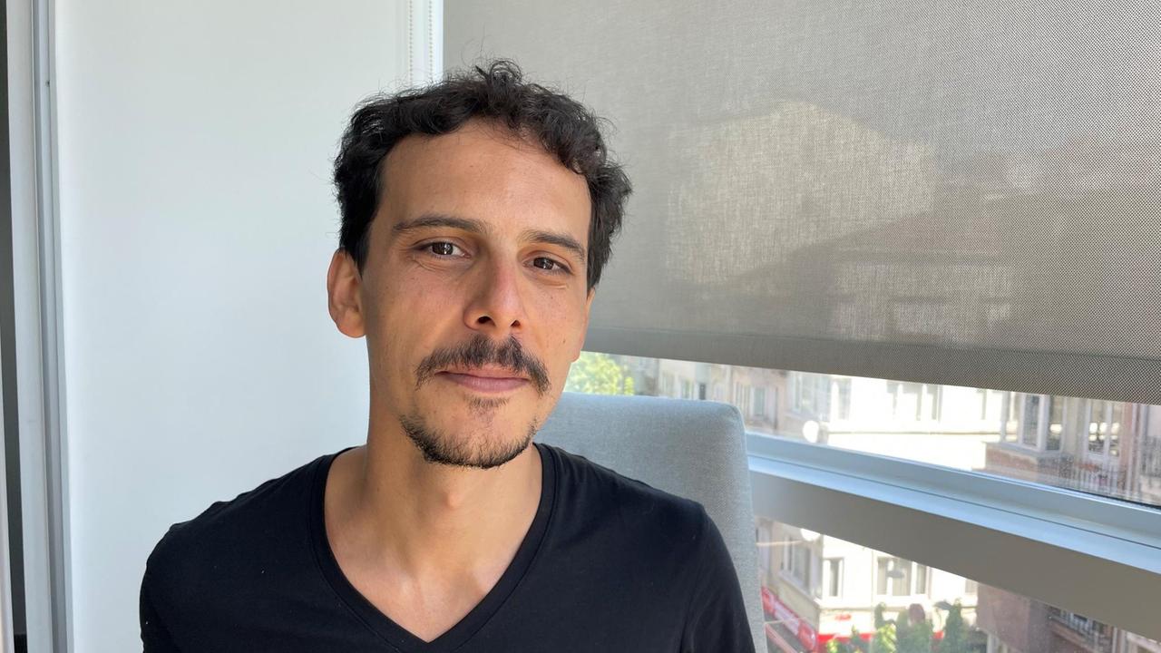 Ein junger Mann mit Bärtchen und dunklen kurzen Haaren sitzt im T-Shirt am Fenster.