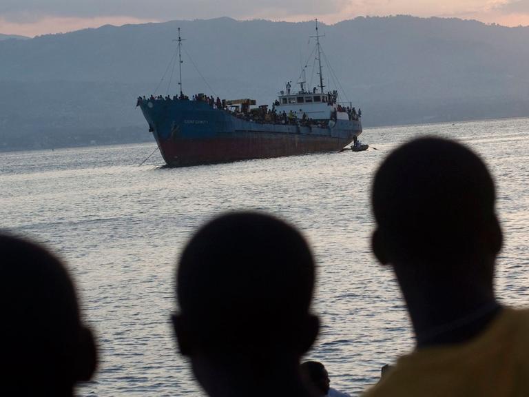 Junge Männer auf Haiti betrachten ein Schiff, auf dem sich viele Flüchtlinge befinden.