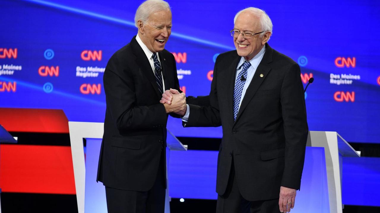 Joe Biden und Bernie Sanders stehen in einer Fernseh-Sendung nebeneinander und reichen sich die Hand.