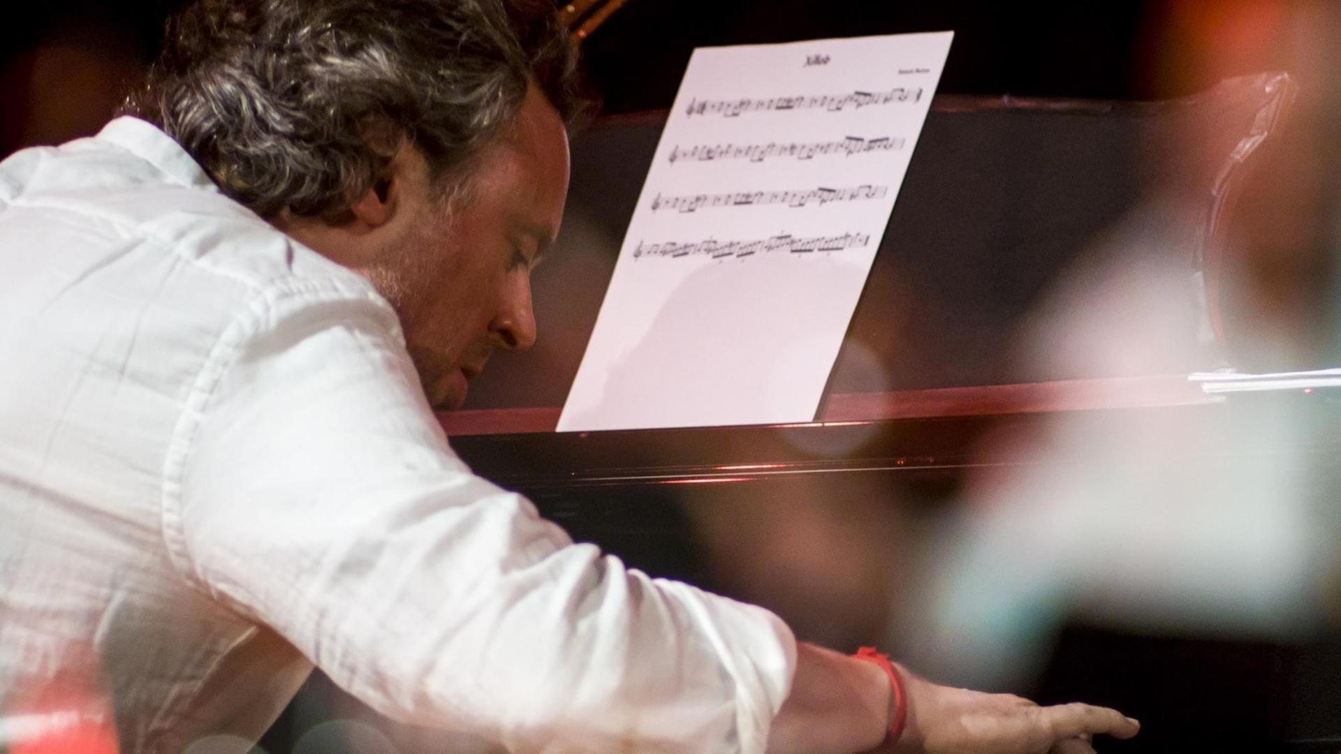 Ein Mann im weißen Hemd spielt mit gesenktem Kopf und gebeugtem Rücken Klavier. Er ist dabei seitlich von hinten zu sehen.