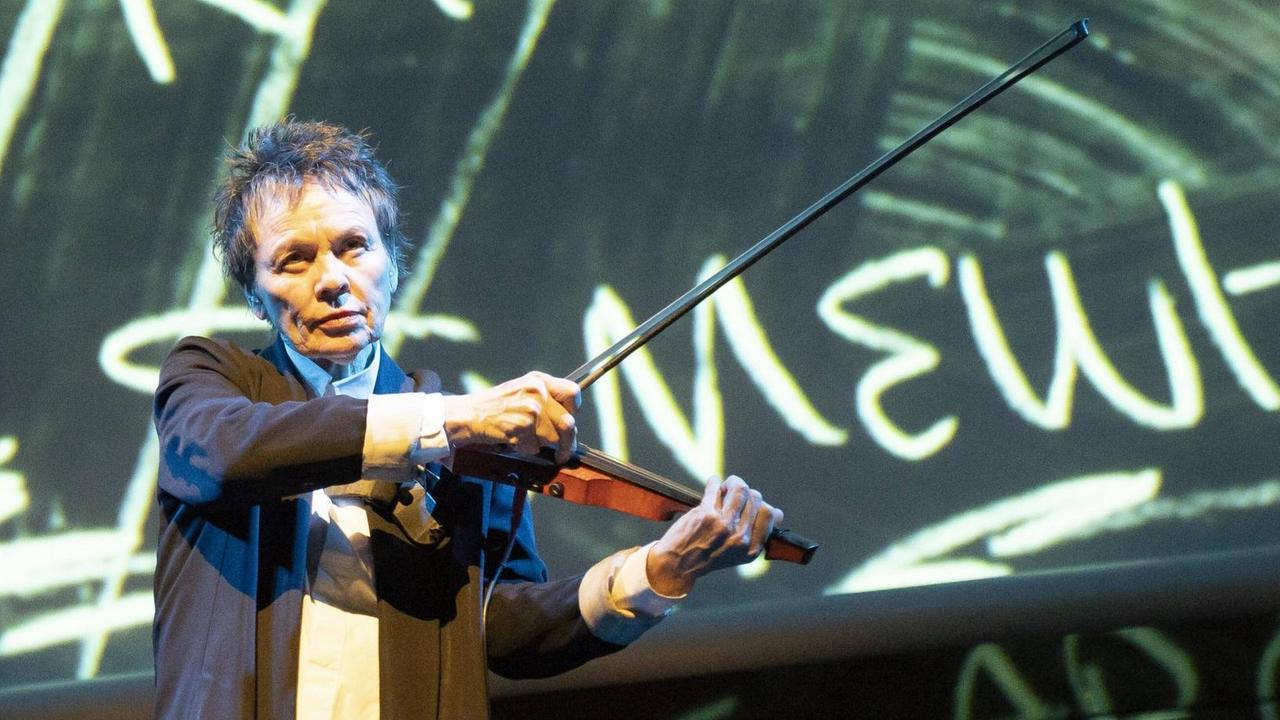 Eine Frau mit kurzen dunkelgrauen Haaren hält eine Violine in ihrer linken Hand und gegen den Oberarm gedrückt. Mit der rechten Hand greift sie an die Saiten, als würde sie sie zupfen. Im Hintergrund ist unscharf ein Schriftzug wie auf einem großen Bildschirm zu erkennen.