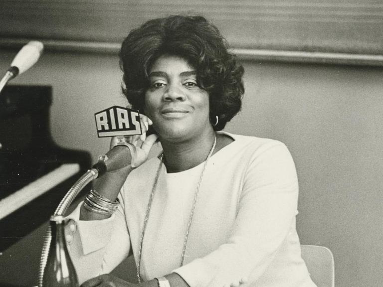 Schwarz-Weiß-Aufnahme der afroamerikanischen Opernsängerin Vera Little in weißer Kleidung vor einem RIAS-Mikrofon.