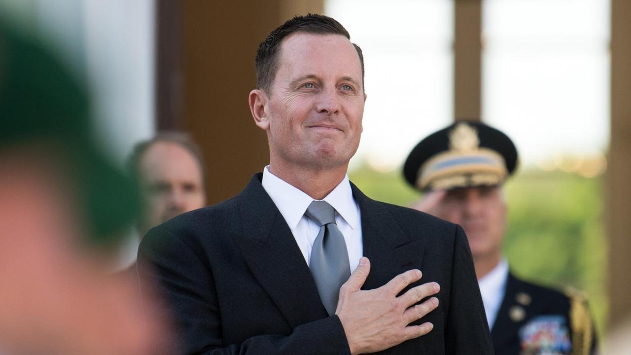 Grenell steht in einem  Raum und hält die rechte Hand ans Herz. Dahinter zwei weitere Männer, einer davon in Uniform. Er salutiert.