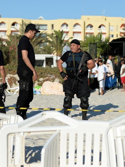 Vier schwarzgekleidete Polizisten stehen einen Tag nach dem Attentat am Strand vor dem Hotel Imperial Marhaba: Die Polizisten tragen Schusswesten. Im Vordergrund sieht man umgestürzte Liegestühle.