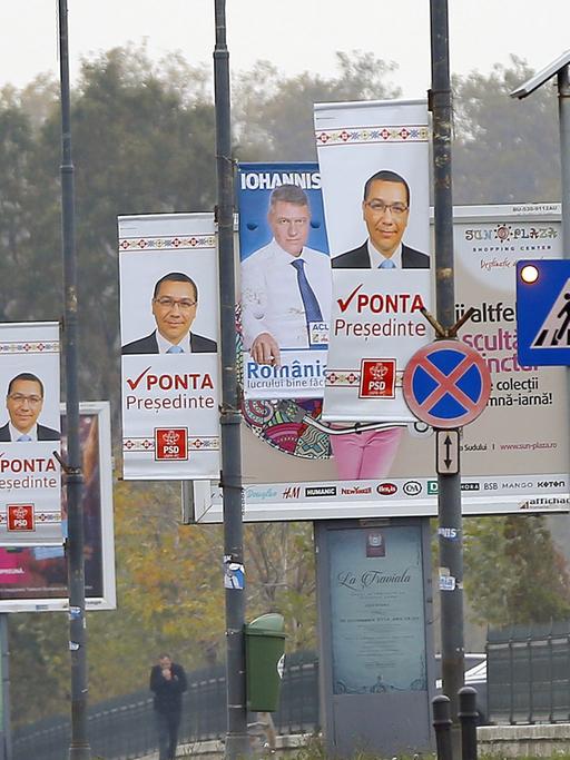 Rümänische Fußgänger passieren eine Reihe Straßenlaternen, an denen Wahlplakate der Bewerber für die rumänische Präsidentschaftswahl hängen.