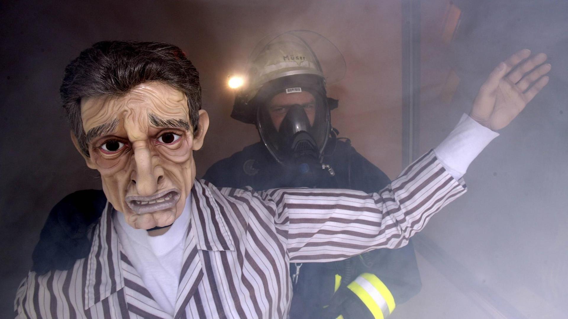 Ein Feuerwehrmann mit Atemschutz und eine lebensgroße Puppe stehen an einem Fenster, in der Luft hängt dichter Rauch