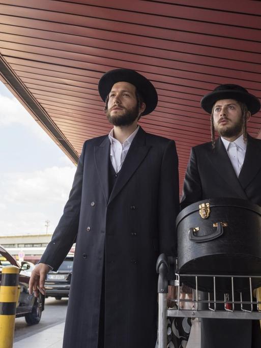 Szene aus der Miniserie "Unorthodox": Zwei ultraorthodoxe Juden laufen mit einem Gepäckwagen vor einem Flughafen entlang.