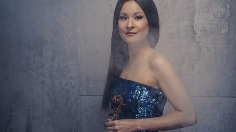 Die Geigerin steht in einem kargen Raum mit ihrer Geige in einem blauben Abendkleid.