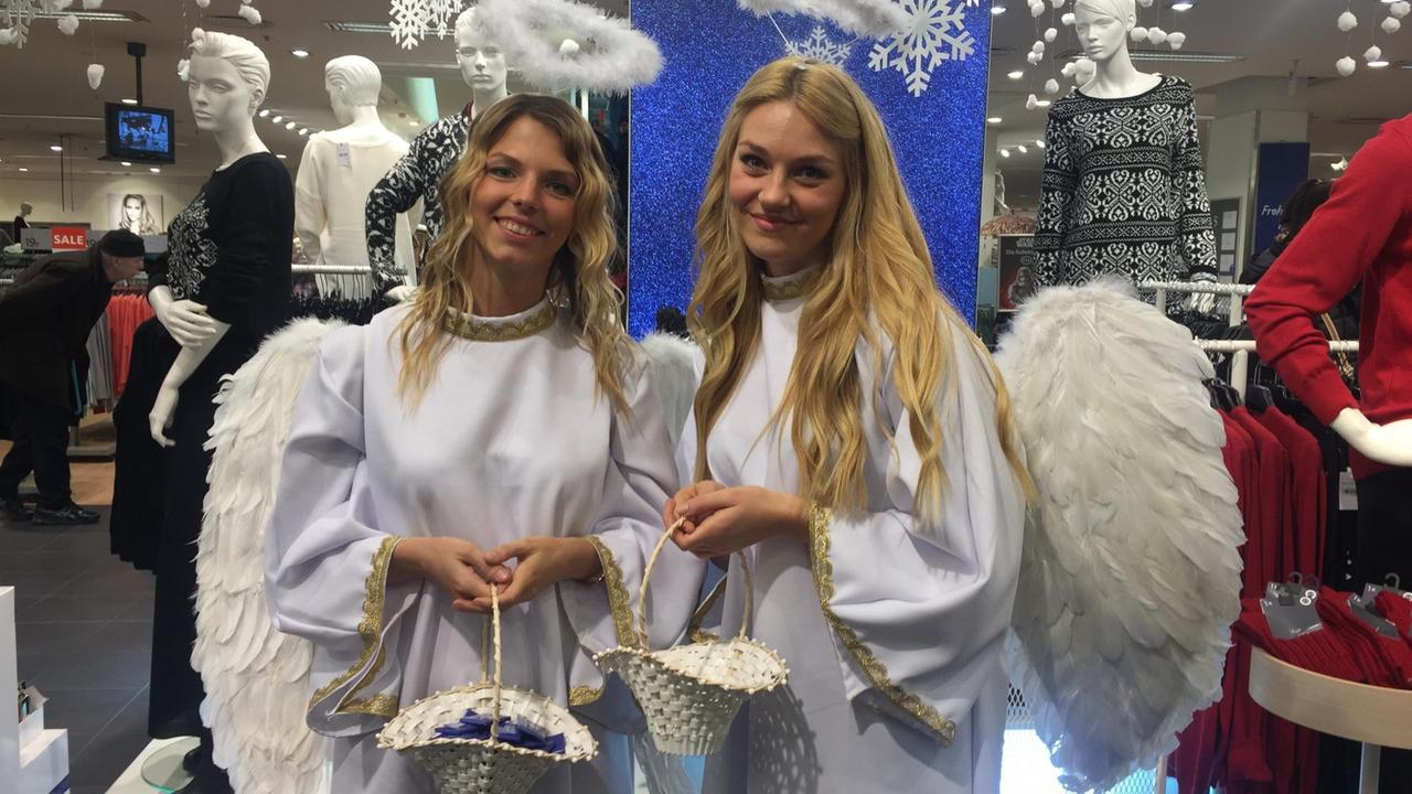 Die Studentinnen Lissi Schneider und Nadine Stührke stehen als Weihnachtsengel verkleidet in einem Kaufhaus vor Schaufensterpuppen. In der Hand halten sie weiße Körbe mit Schokoladentäfelchen für die Besucher.
