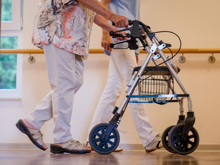 Eine Altenpflegerin begleitet eine ältere Frau mit dem Rollator durch den Flur eines Pflegeheims.