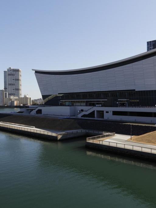 Blick auf eine Wasserfläche an deren Ufer eine moderne Sportarena steht — im Hintegrund sind Hochhäuser zu sehen