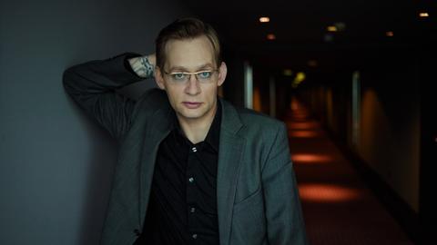 Clemens Meyer lehnt in einem dunklen Hotelflur an einer Wand und schaut in die Kamera.