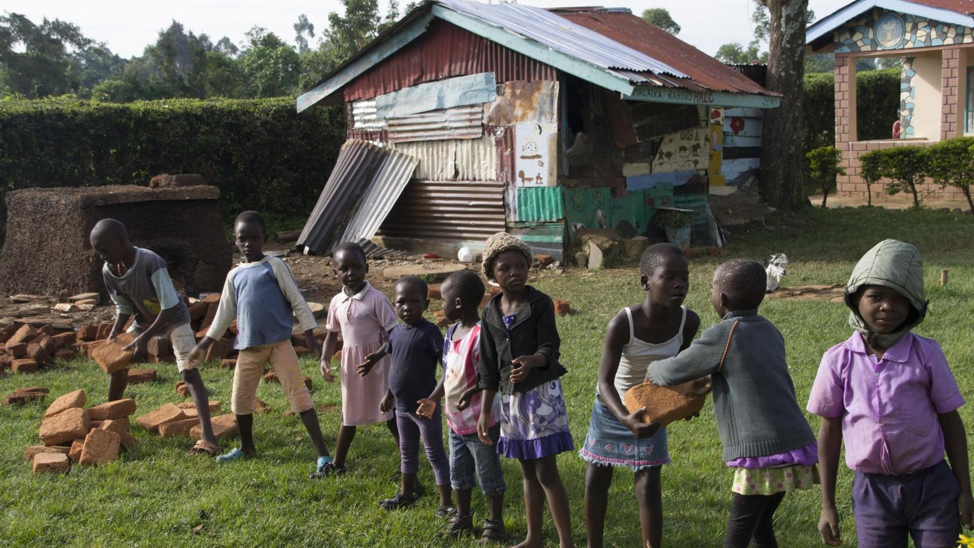 Kleine afrikanische Kinder haben sich in einer Reihe aufgestellt und bilden eine Kette, mittels derer Ziegelsteine für den Bau der Mädchentoiletten transportiert werden.