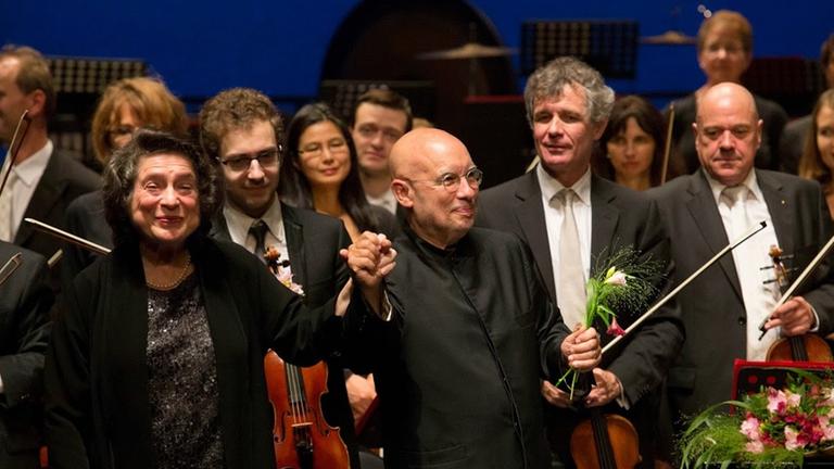 Die Pianistin und ein Dirigent erhalten vor dem Orchester stehend Beifall. Blumen liegen auf dem Flügel.