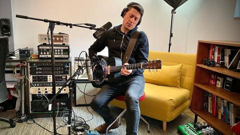Ein Mann mit schwarzen kurzen Haaren sitzt auf einem Hocker vor Mikrofonen und spielt Gitarre. Er trägt Kopfhörer, hinter ihm steht ein gelber Sessel, ein Regal und technisches Equipment.