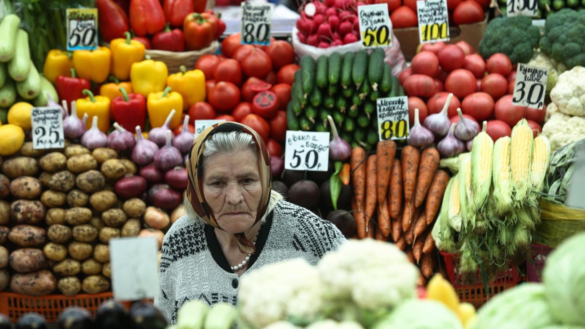 Ein Stand mit Gemüse und einer älteren Verkäuferin mit Kopftuch auf dem Domodedovsky Lebensmittel Markt in Moskau, Russland