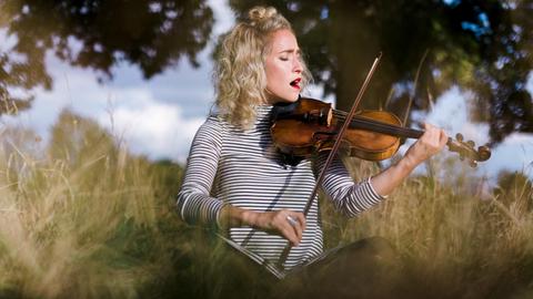 Die Geigerin Julia Lacherstorfer spielt Geige sitzend auf einer Wiese im Freien unter Bäumen