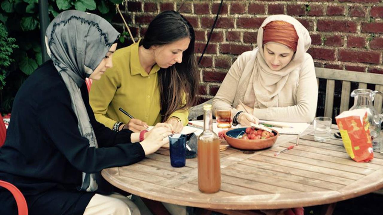 Zwei Frauen mit Kopftuch und eine weitere junge Frau sitzen an einem Gartentisch und arbeiten.