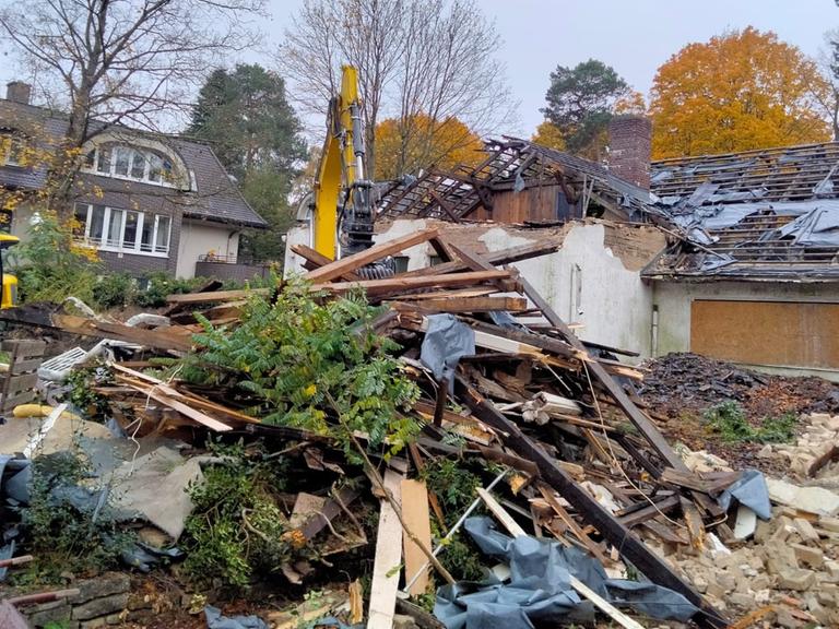 Ein Bagger steht hinter einem Schutthaufen vor einem halb abgerissenem Haus.