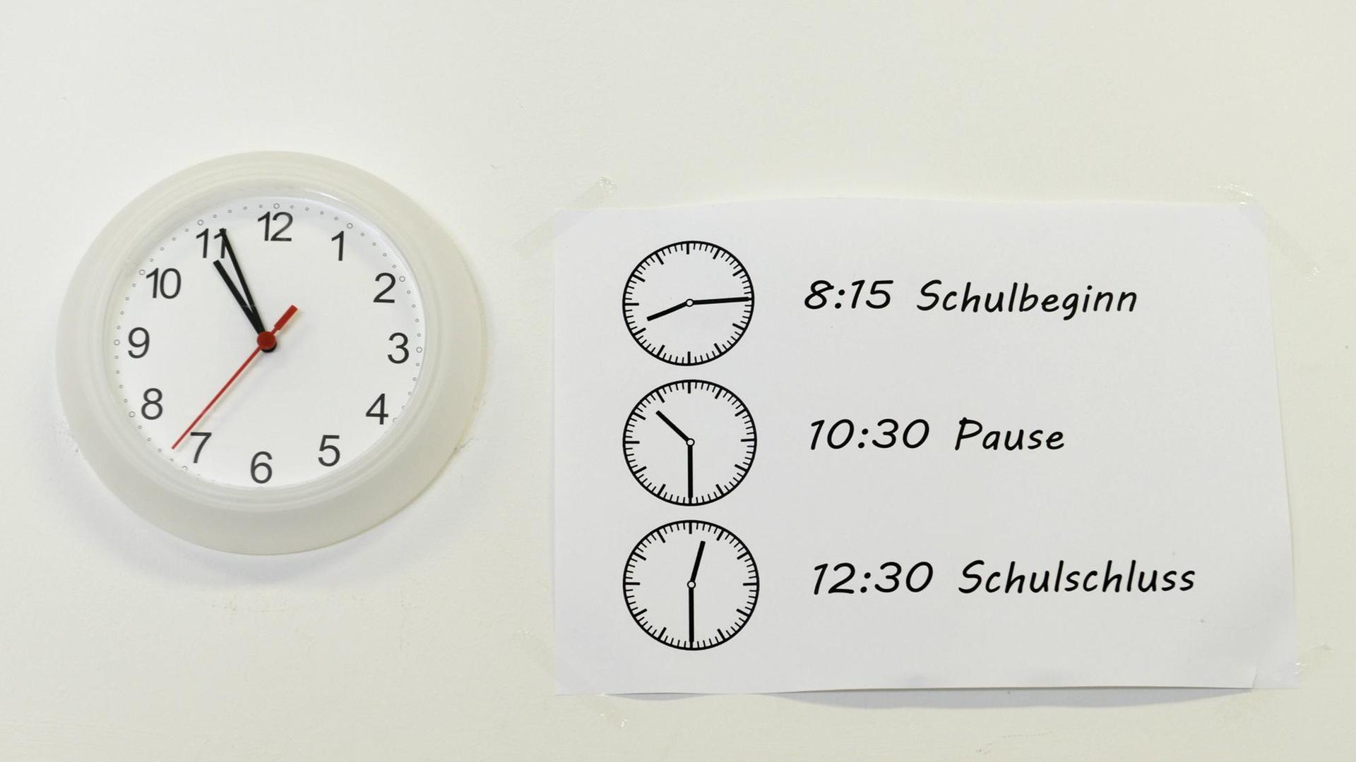 Schulstart- und Pausenzeiten stehen auf einem Zettel, der neben einer Uhr hängt.