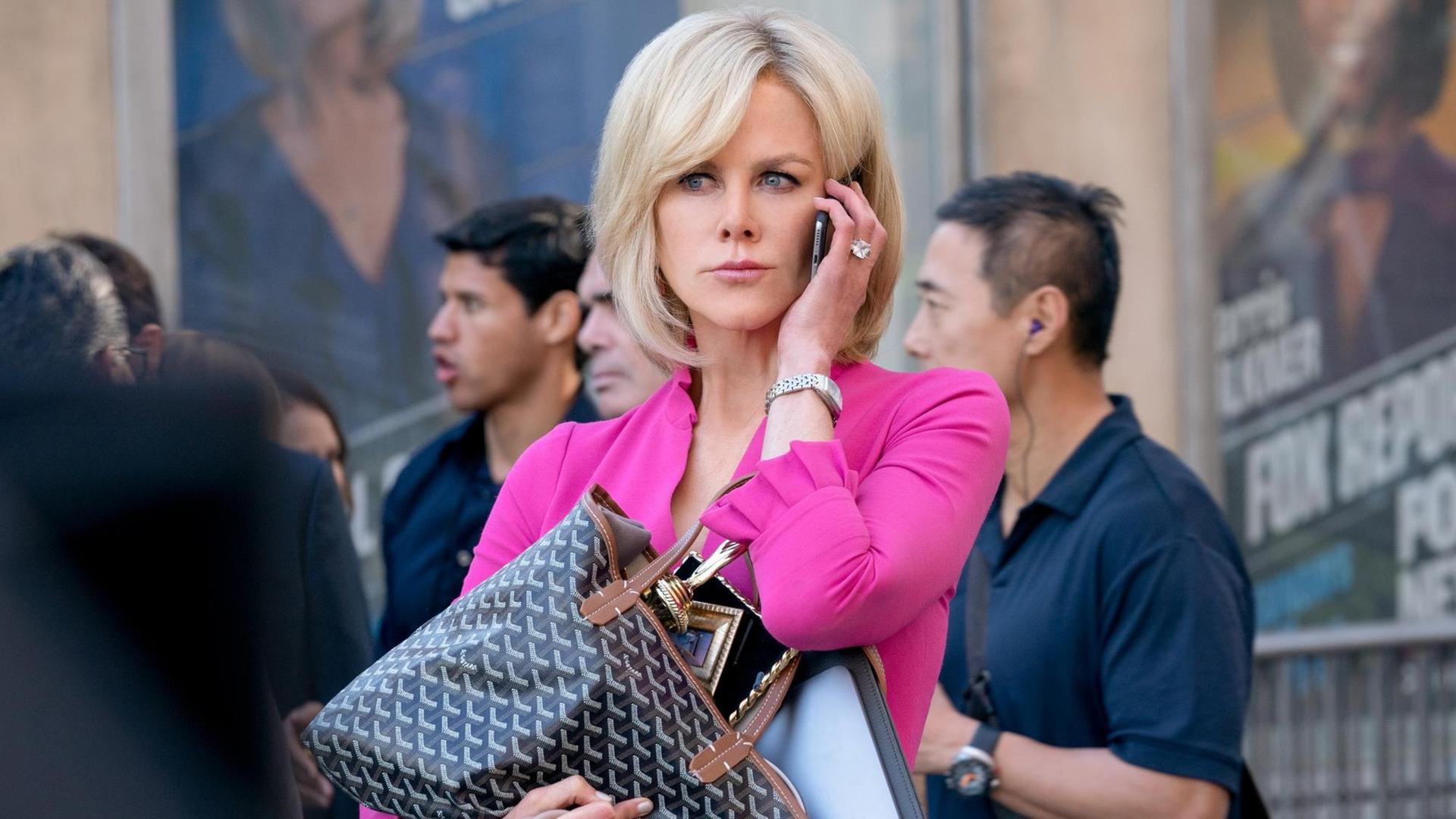 Szene Nicole Kidman als Gretchen Carlson in "Bombshell": Sie steht mit sehr blonden Haaren in einem pinken Kostüm vor einem Kino, um sie herum Menschen. Sie hält sich ein Smartphone ans Ohr und hält mit der anderen Hand eine prall gefüllte große Handtasche.