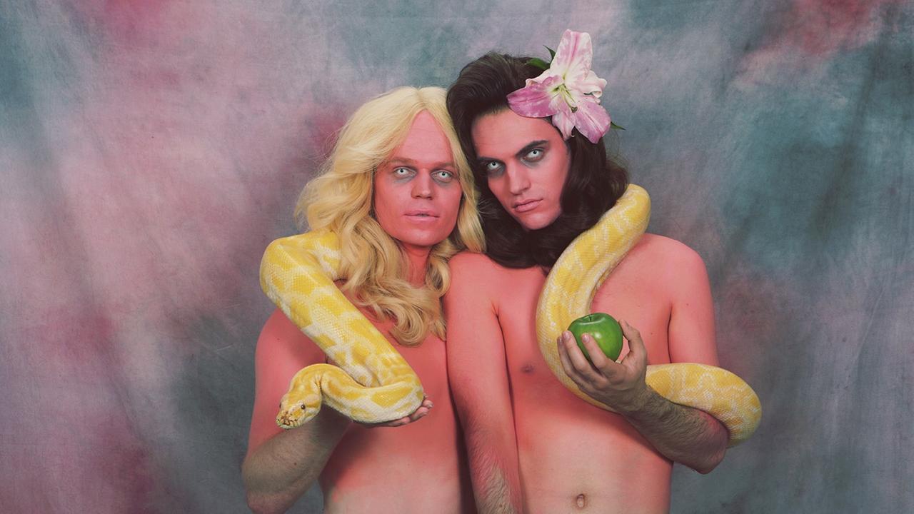 Sam Dust (rechts) und Connan Mockasin, vom walisisch-australischen Duo Soft Hair, blicken in die Kamera. Sie sind mit nacktem Oberköper abgebildet, tragen lange Perücken und eine große, gelbe Schlange um den Hals. Dust hat eine Blume im Haar und hält einen grünen Apfel in der Hand.