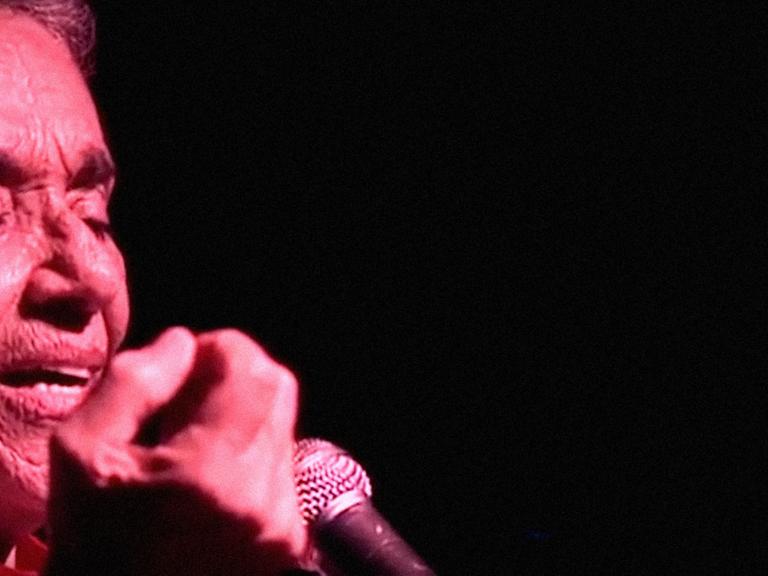 Nahaufnahme von Chavela Vargas während eines Konzertes: Sie singt mit geschlossenen Augen und wird von rotem Licht angestrahlt, der Hintergrund ist dunkel.
