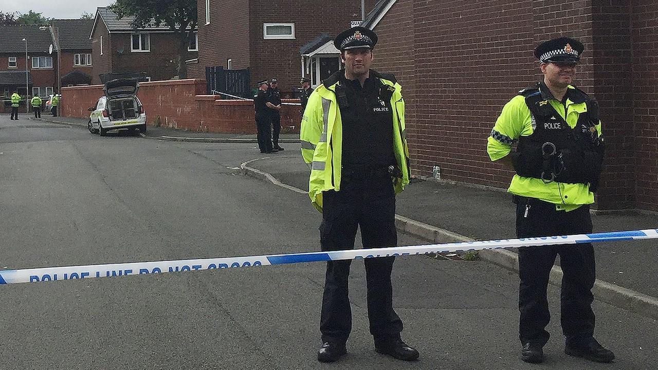 Zwei Polizisten in leuchtgelben Warnwesten stehen hinter Absperrband auf einer Straße mit englischen Reihenhäusern aus Backstein. Im Hintergrund weitere Polizisten und ein Polizeiauto. Zwei Polizisten in leuchtgelben Warnwesten stehen hinter Absperrband auf einer Straße mit englischen Reihenhäusern aus Backstein. Im Hintergrund weitere Polizisten und ein Polizeiauto.