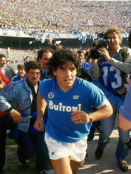 Diego Maradona läuft durch das Stadium, gefolgt von Journalisten.