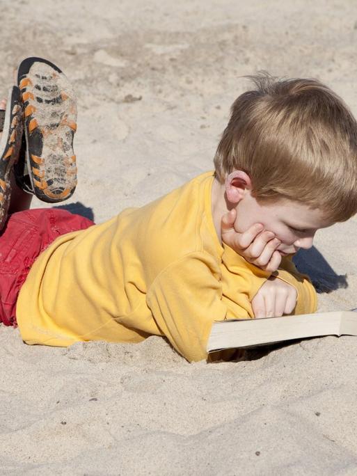 Ein kleiner Junge liegt am Strand und liest vertieft in einem Buch. (Quelle: imago)