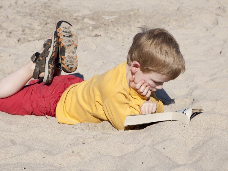 Ein kleiner Junge liegt am Strand und liest vertieft in einem Buch. (Quelle: imago)