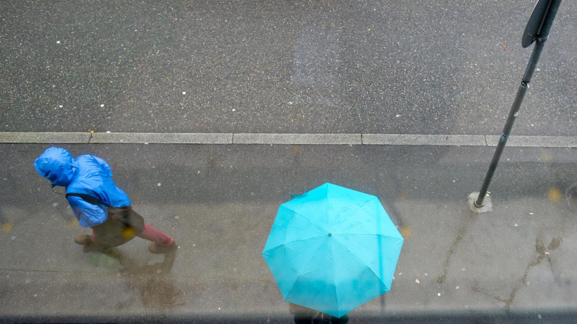 Zwei Passanten laufen bei Nieselregen mit einem türkisfarbenem Regenschirm und blauer Regenjacke den Bürgersteig entlang.