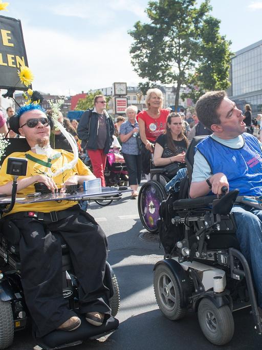 Drei Demonstranten bei der "Pride Parade 2014" tragen Transparente und Schilder. Auf einem Schild steht: "Bitte auffallen!". Zwei Demonstranten fahren in Rollstühlen, einer läuft. Im Hintergrund weitere Demonstranten.