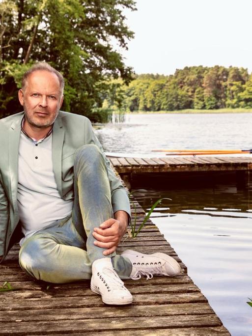 Axel Milberg sitzt bekleidet mit Jeans, Sakko und Turschuhen auf einem Bootssteg. Im Hintergrund ein See und Ruderboote.