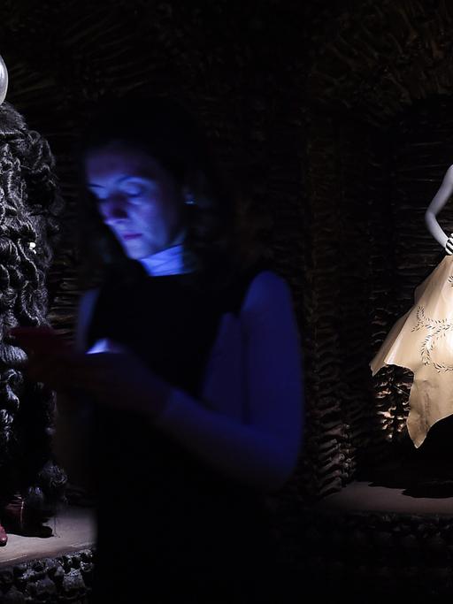 Zwei Kleiderpuppen, eine in einem weißen Kleid, eine in einem schwarzen Kleid, beleuchtet in einem dunklen Ausstellungsraum, im Vordergrund ist schwach eine Besucherin zu sehen.