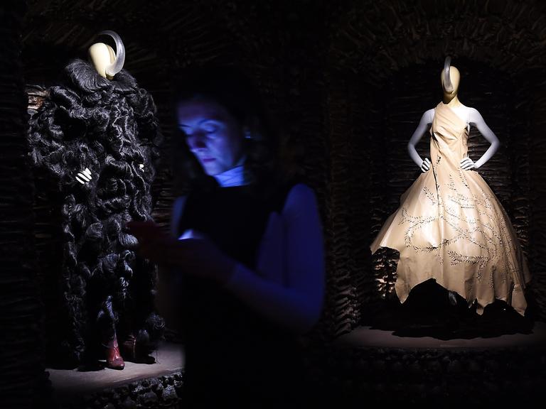 Zwei Kleiderpuppen, eine in einem weißen Kleid, eine in einem schwarzen Kleid, beleuchtet in einem dunklen Ausstellungsraum, im Vordergrund ist schwach eine Besucherin zu sehen.