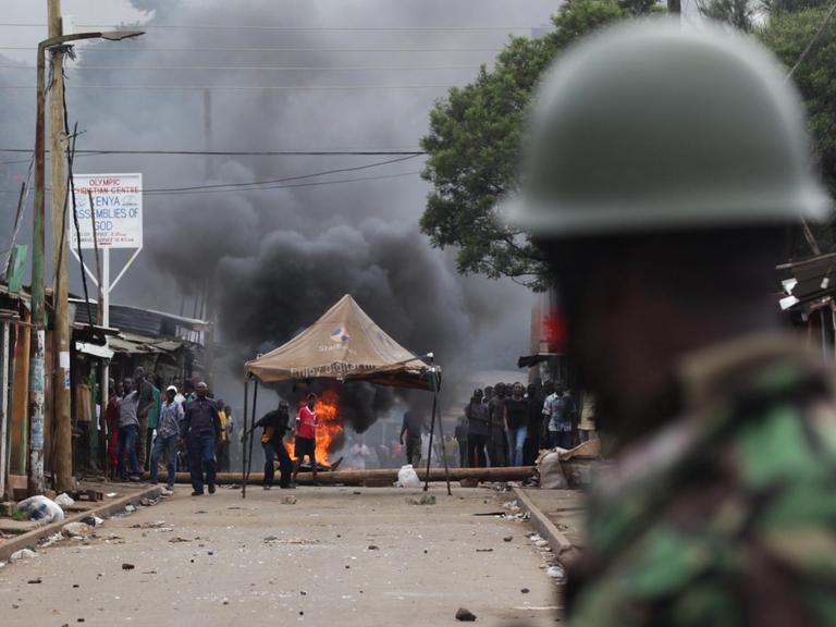 Ein Polizist blickt auf brennende Barikaden und Demonstranten in Kibera, Nairobi