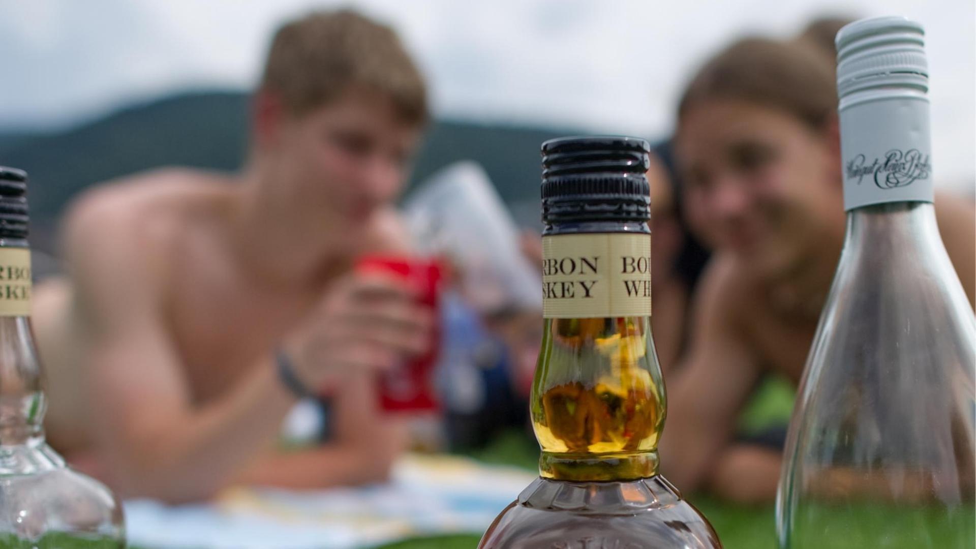 Auf dem Bild sieht man zwei Jugendliche auf einer Wiese, sie trinken Alkohol. Im Vordergrund stehen zwei Flaschen mit Alkohol.