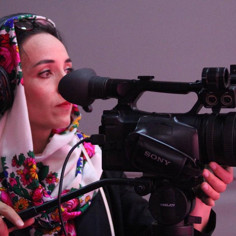 Eine Frau steht an einer TV-Kamera und filmt. Sie trägt ein geblümtes Kopftuch.