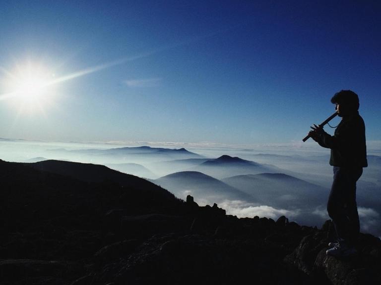 Symbolbild Musik und Natur: Silhouette eines Mannes, der in einer Gebirgslandschaft auf einem Gipfel Flöte spielt.