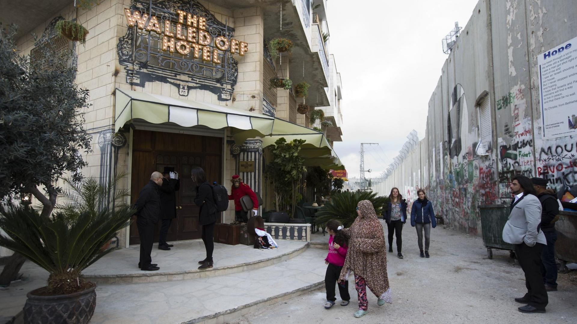Passanten gehen in Bethlehem (Westjordanland) am Eingangsbereich des "The Walled Off Hotel" und israelischen Sperranlagen vorüber. Das Hotel in der Nähe der israelischen Sperranlagen zeigt Motive des Künstlers Banksy und wirbt mit dem "schlechtesten Blick der Welt".
