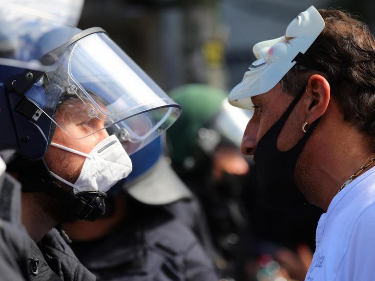 Ein Mann mit Mundschutz unter der Nase und hochgeschobener Anonymous-Maske schreit einen Polizisten an.