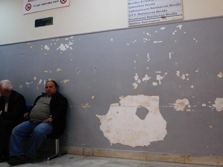 Männer sitzen auf Stühlen vor einer Wand mit abgeplatzter Farbe und Putz in einem Krankenhaus und warten.