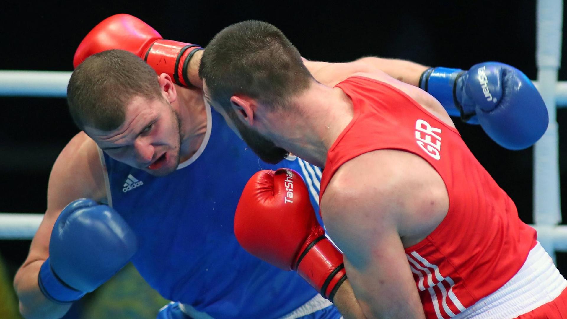 Ein in blau und ein in rot gekleideter Boxer kämpfen in einem Ring; der rote schlägt knapp am Kopf des blauen vorbei. Dahinter die weißen Seile des Rings vor einem dunklen Hintergrund.